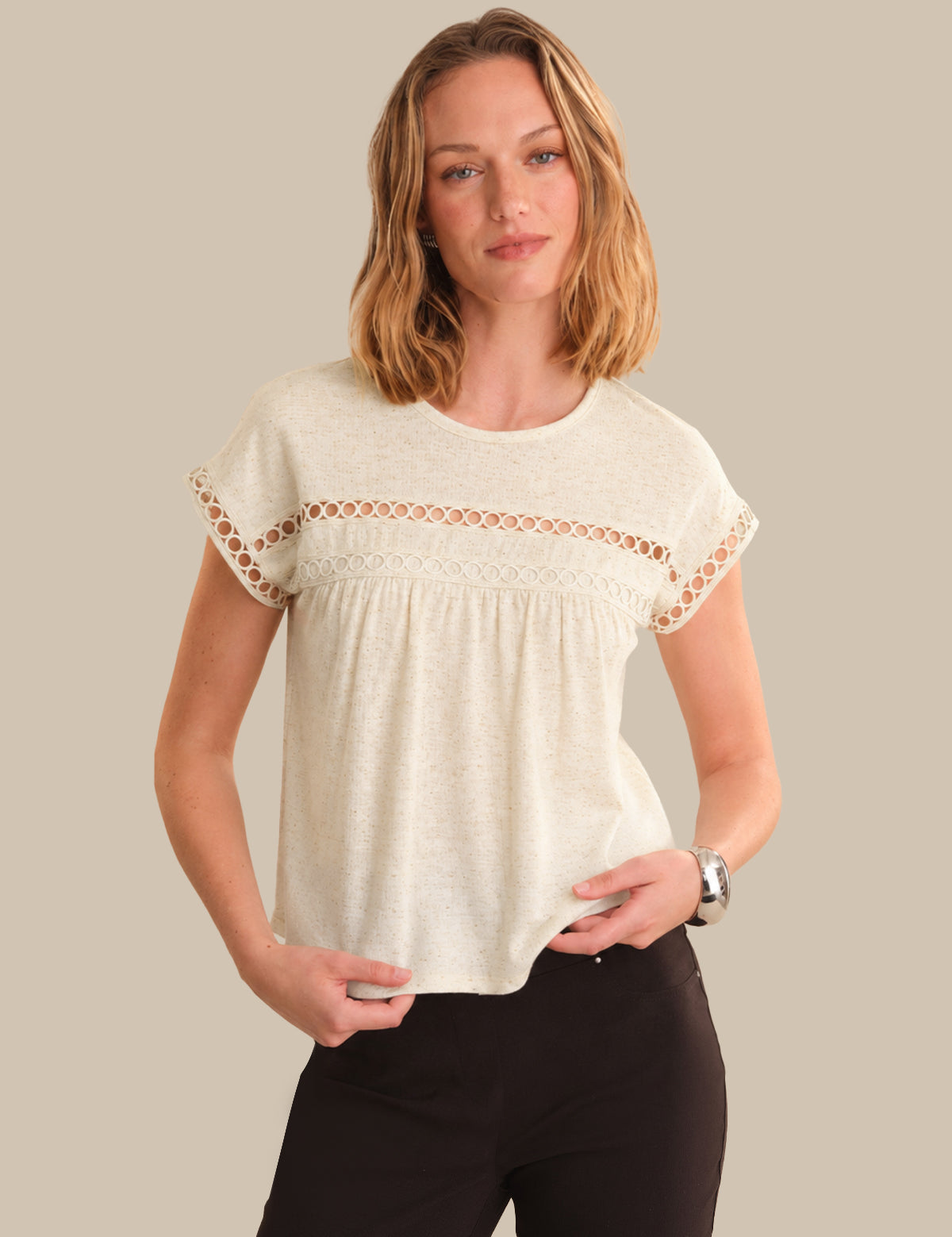 A woman modeling a short-sleeve cream-colored top with a decorative lace yoke and scalloped edges, paired with black pants, against a solid beige background.