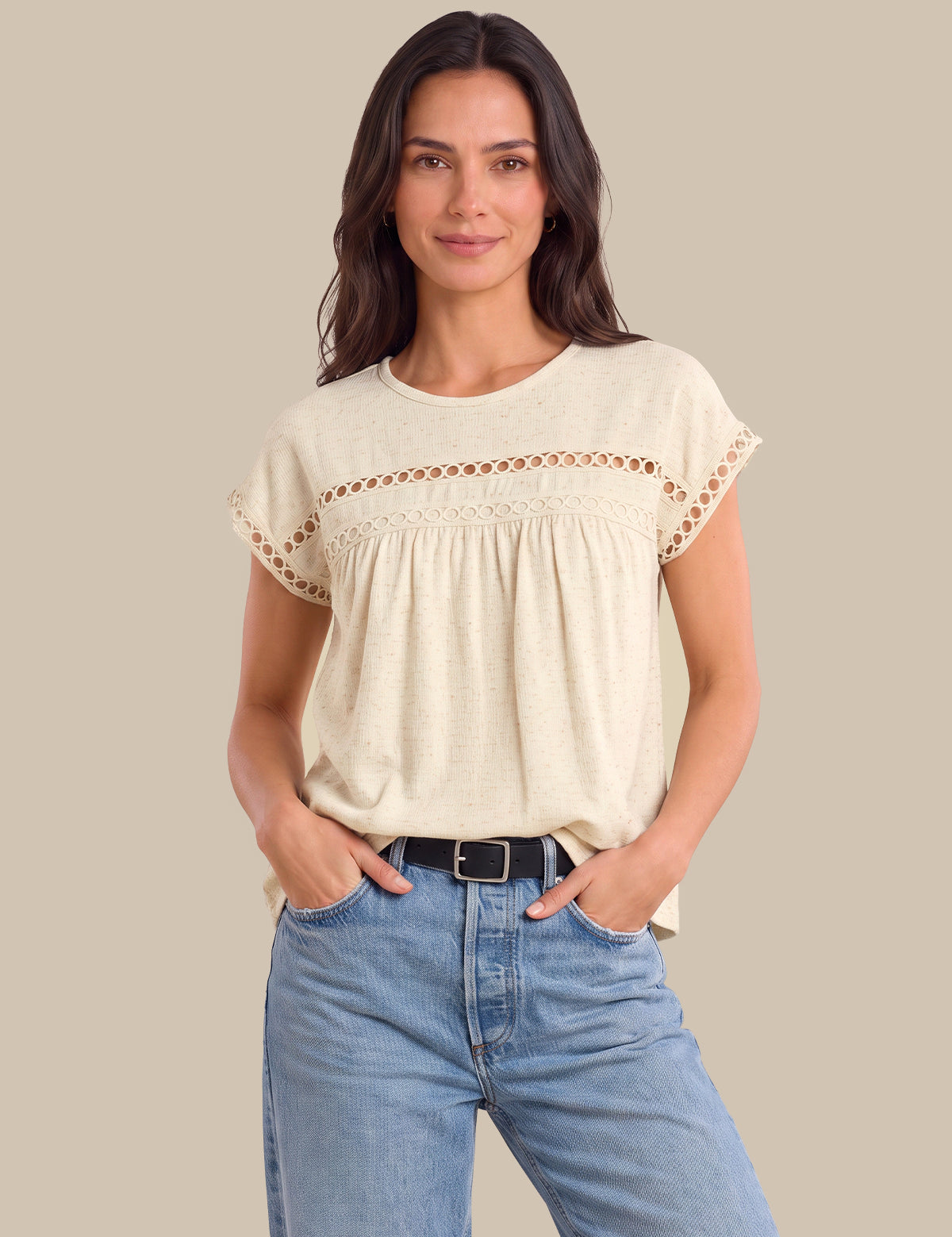 A woman modeling a cream-colored short-sleeve top with delicate crochet lace detailing across the chest and sleeve cuffs, paired with light blue jeans against a solid beige background.