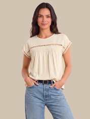 A woman modeling a cream-colored short-sleeve top with delicate crochet lace detailing across the chest and sleeve cuffs, paired with light blue jeans against a solid beige background.
