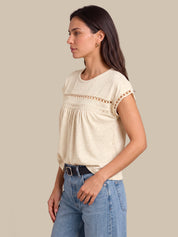A woman wearing a cream-colored short-sleeve top with a decorative crochet lace band across the chest and light blue jeans, shown in a side profile against a solid beige background.