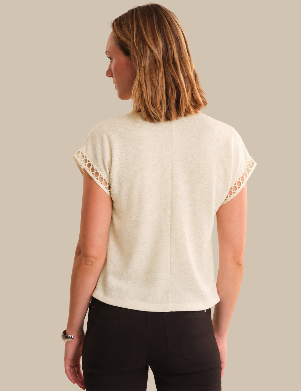 A woman shown from the back wearing a cream-colored knit top with decorative lace trim on the short sleeves, paired with black pants, against a solid beige background.