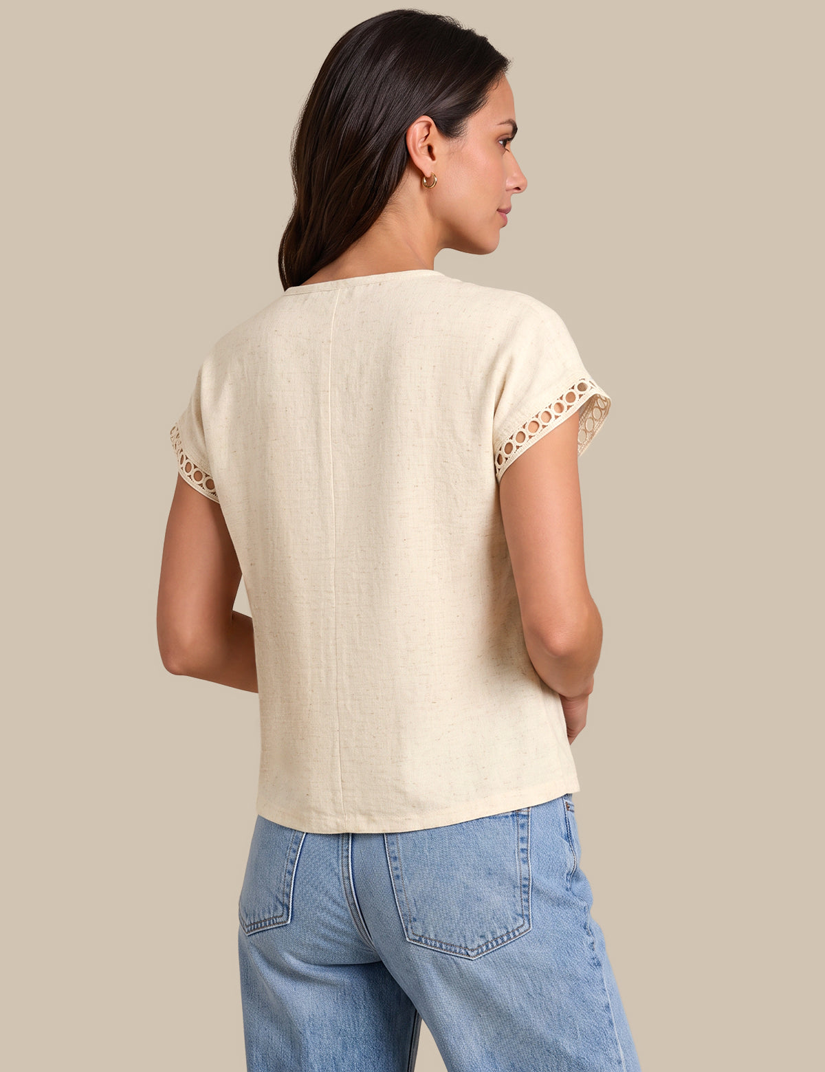 A woman shown from the back wearing a light beige linen short-sleeve top with lace trim at the sleeve cuffs, paired with light blue jeans, against a solid beige background.