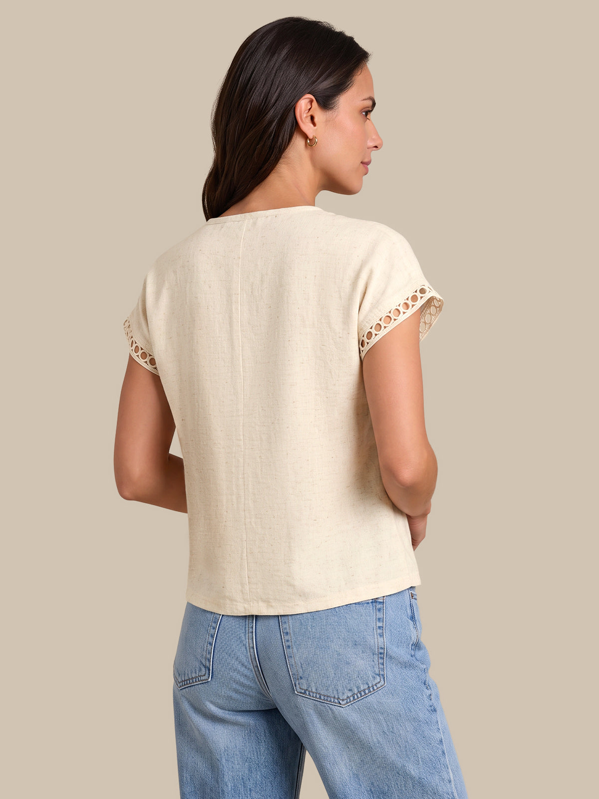 A woman shown from the back wearing a light beige linen short-sleeve top with lace trim at the sleeve cuffs, paired with light blue jeans, against a solid beige background.