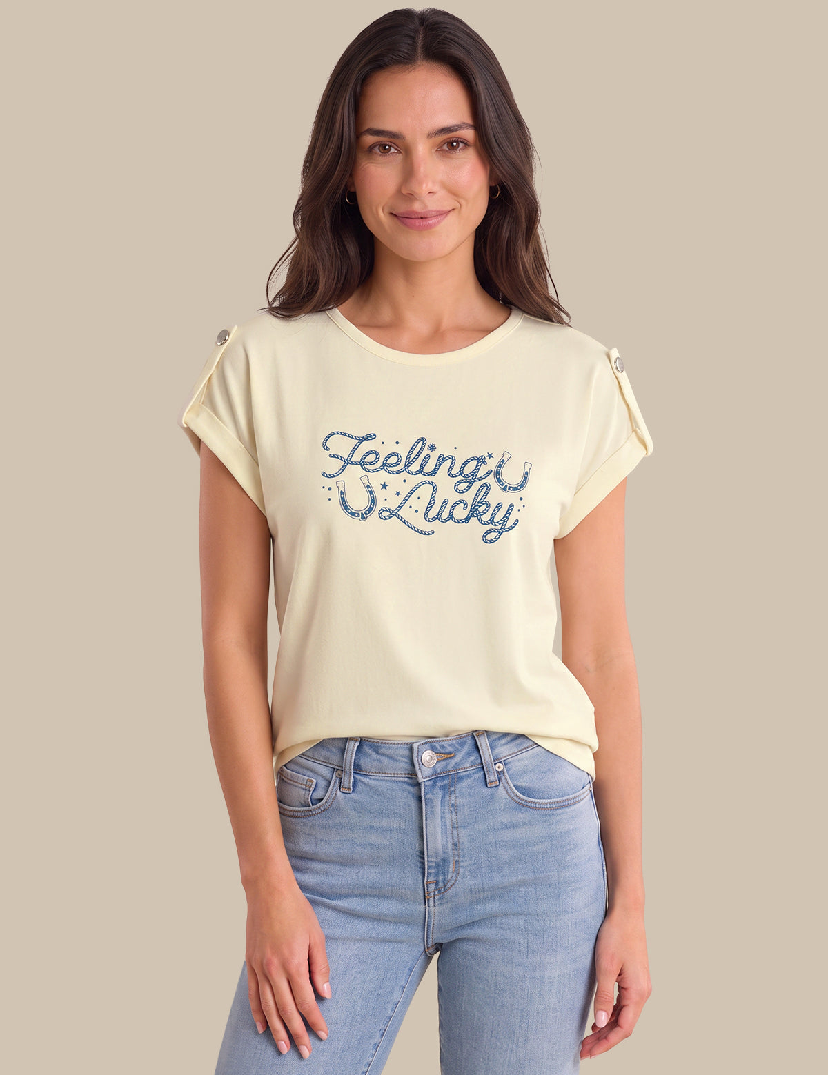A woman modeling a cream-colored short-sleeve t-shirt with 'Feeling Lucky' printed in blue script across the chest, paired with light blue jeans against a solid beige background.