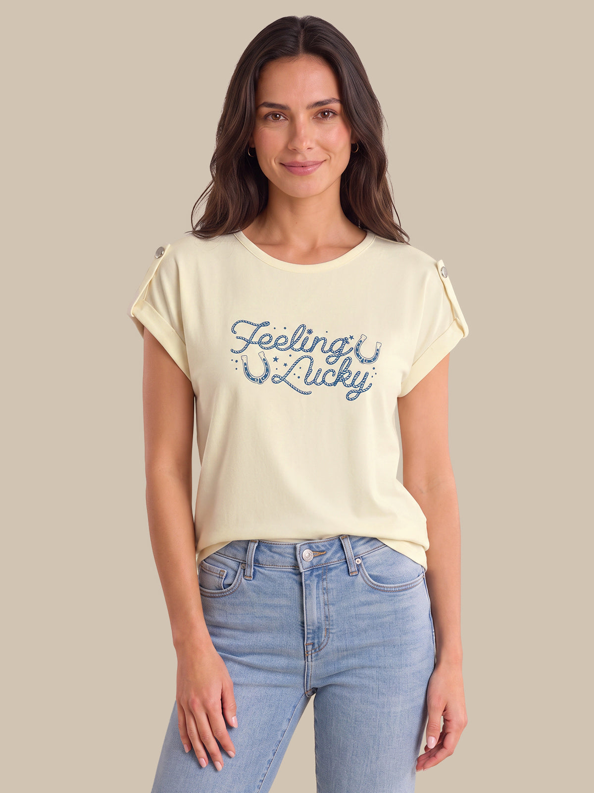 A woman modeling a cream-colored short-sleeve t-shirt with 'Feeling Lucky' printed in blue script across the chest, paired with light blue jeans against a solid beige background.