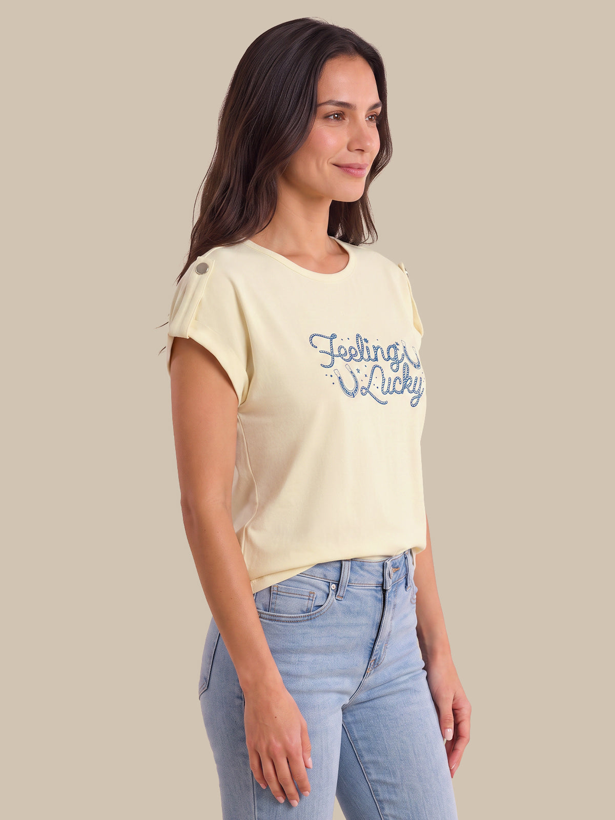 A woman wearing a cream-colored short-sleeve crop top with blue 'Feeling Lucky' text on the front, paired with light blue jeans, set against a solid beige background.