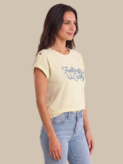 A woman wearing a cream-colored short-sleeve crop top with blue 'Feeling Lucky' text on the front, paired with light blue jeans, set against a solid beige background.