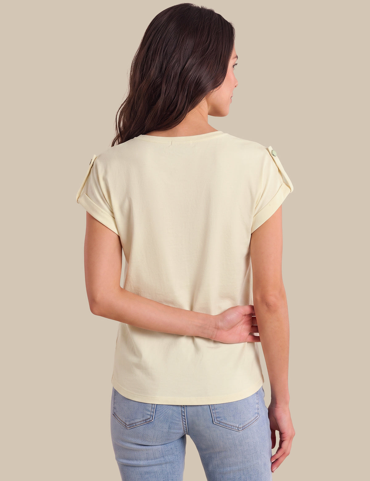 A woman viewed from the back wearing a light cream short-sleeve top with decorative ruffle details on the shoulders and light blue jeans, set against a solid beige background.