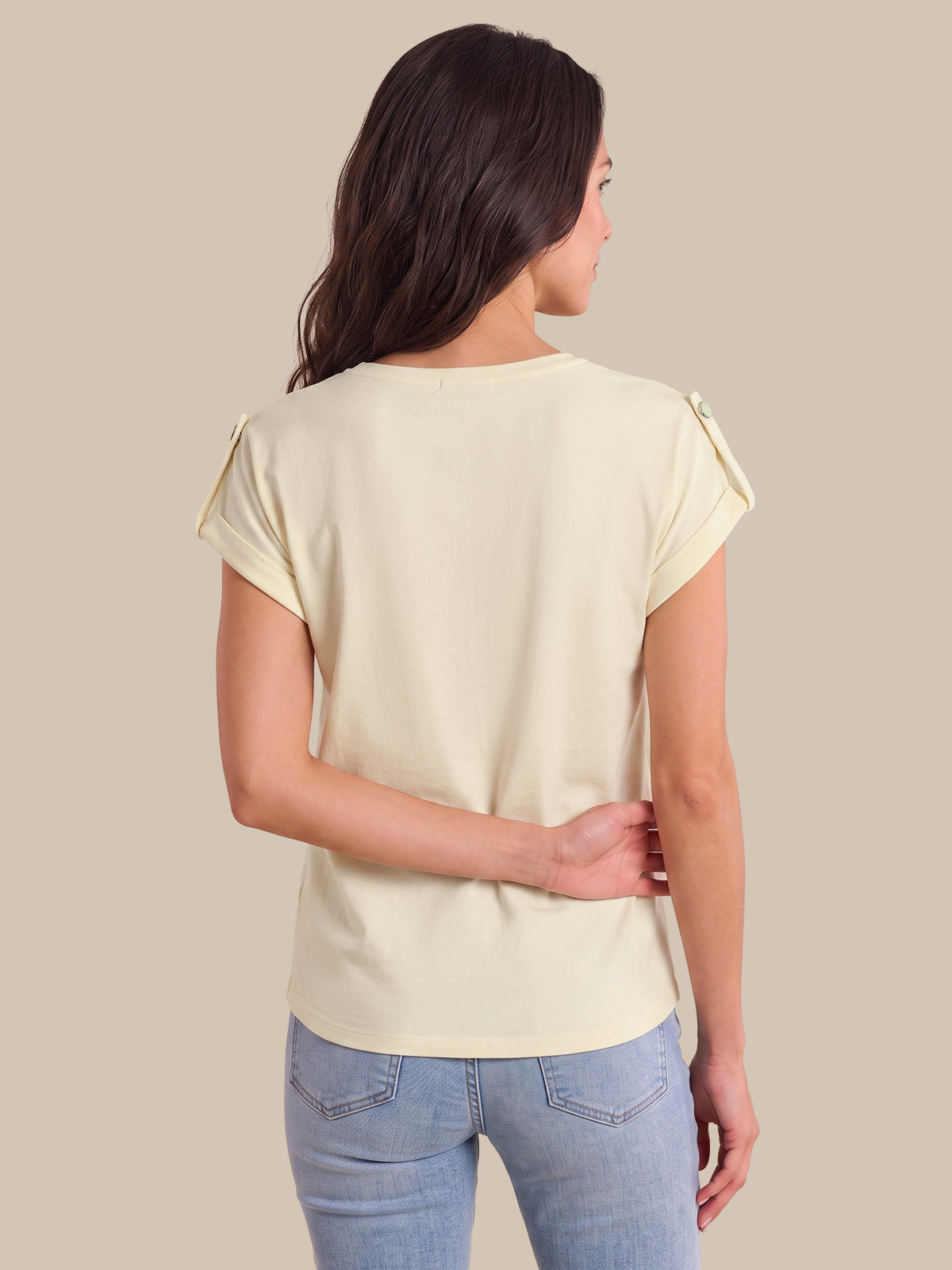A woman viewed from the back wearing a light cream short-sleeve top with decorative ruffle details on the shoulders and light blue jeans, set against a solid beige background.