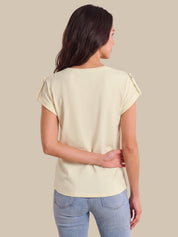 A woman viewed from the back wearing a light cream short-sleeve top with decorative ruffle details on the shoulders and light blue jeans, set against a solid beige background.