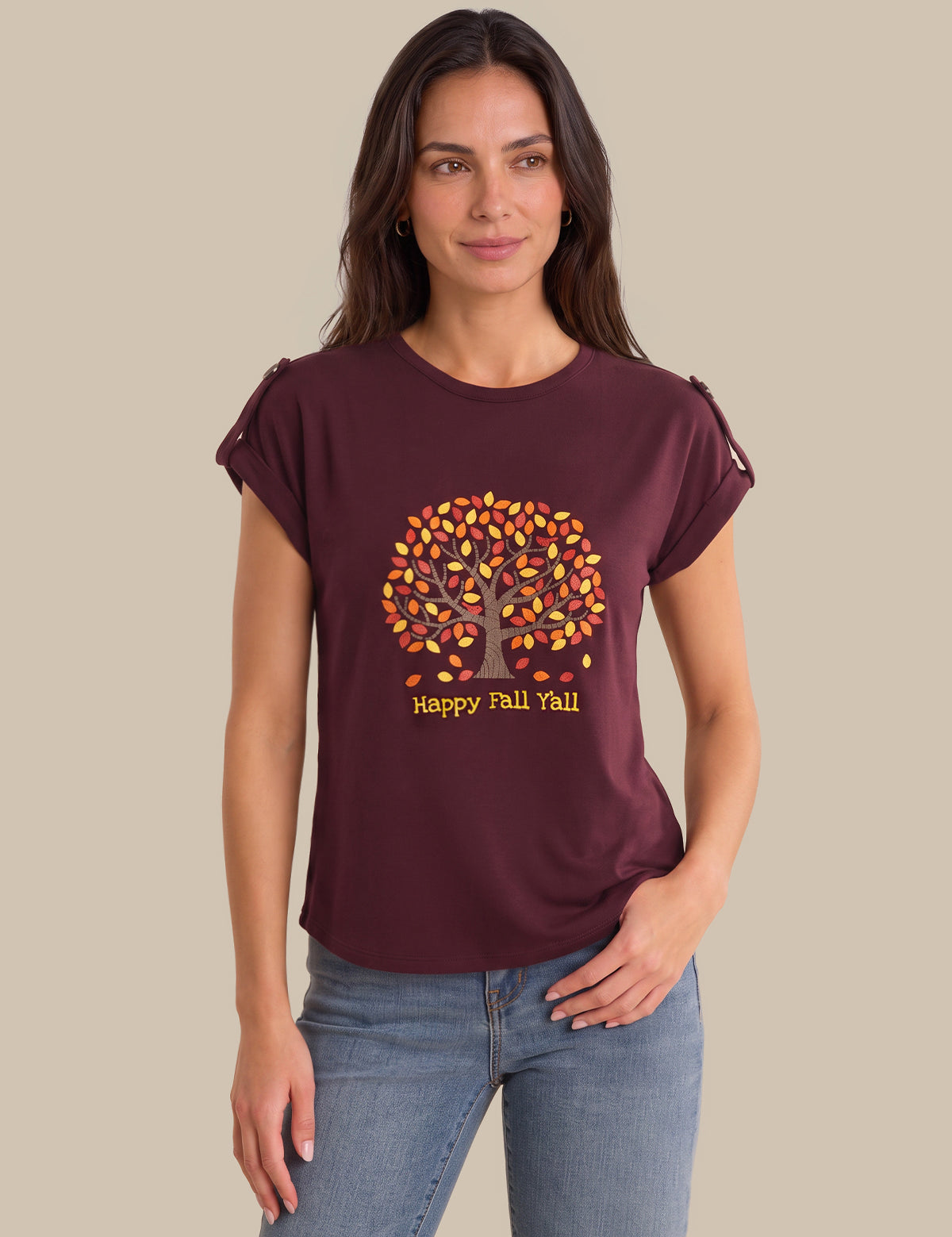 A woman modeling a maroon short-sleeve t-shirt with a 'Happy Fall You!' autumn tree graphic on the front, paired with blue jeans against a solid beige background.