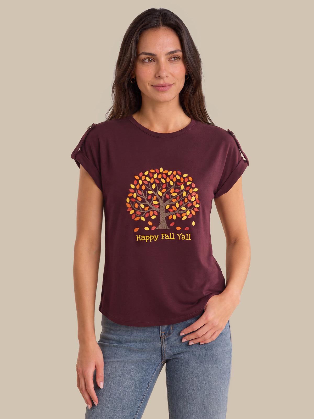 A woman modeling a maroon short-sleeve t-shirt with a 'Happy Fall You!' autumn tree graphic on the front, paired with blue jeans against a solid beige background.