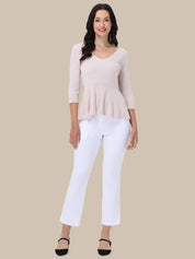 A woman modeling a light pink ribbed knit top with three-quarter sleeves and a peplum hem, paired with white cropped pants and black flat shoes, against a solid beige background.