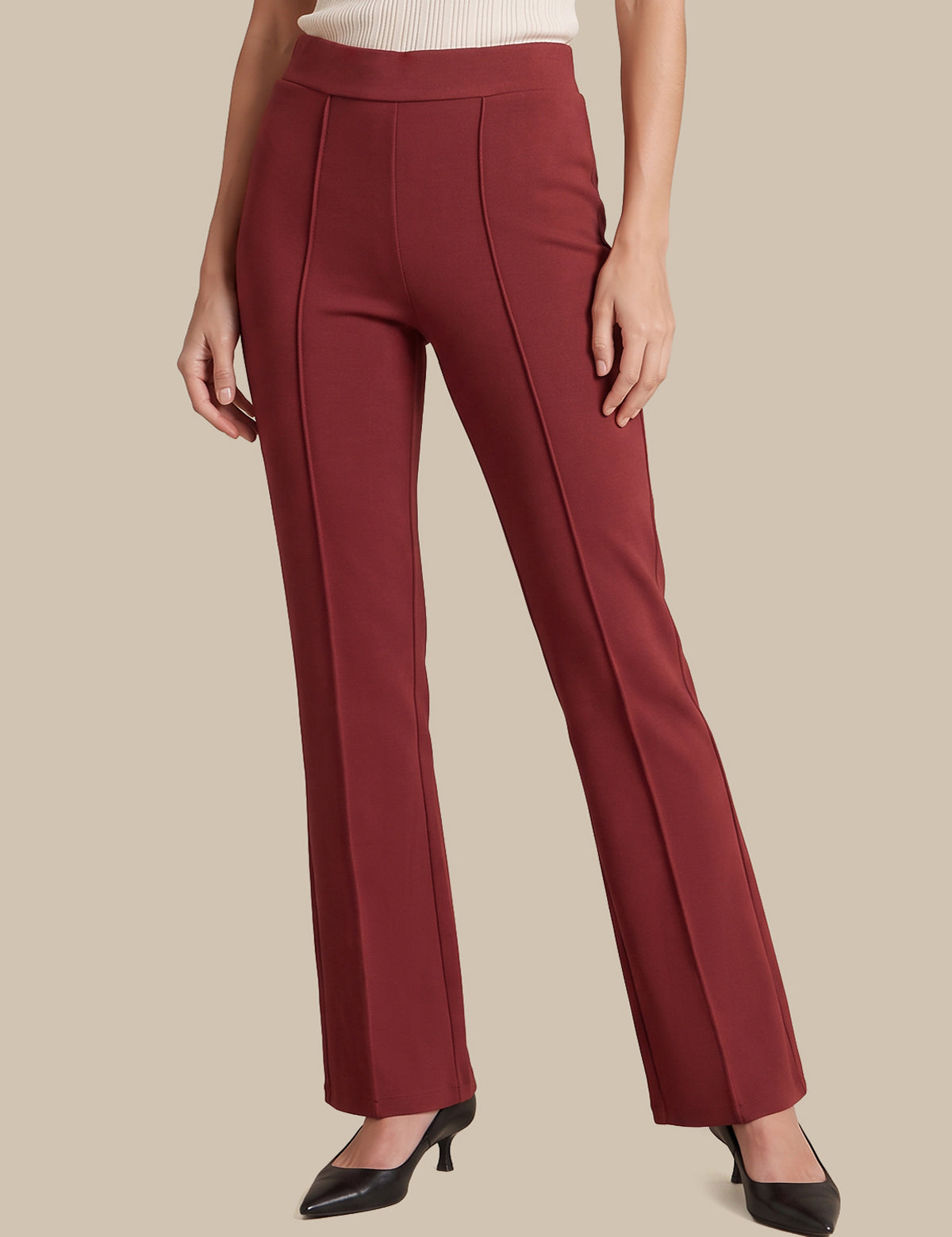 A model wearing a pair of high-waisted, straight-leg trousers in a deep burgundy red, paired with a cream ribbed top and black pointed-toe heels, set against a solid beige background.