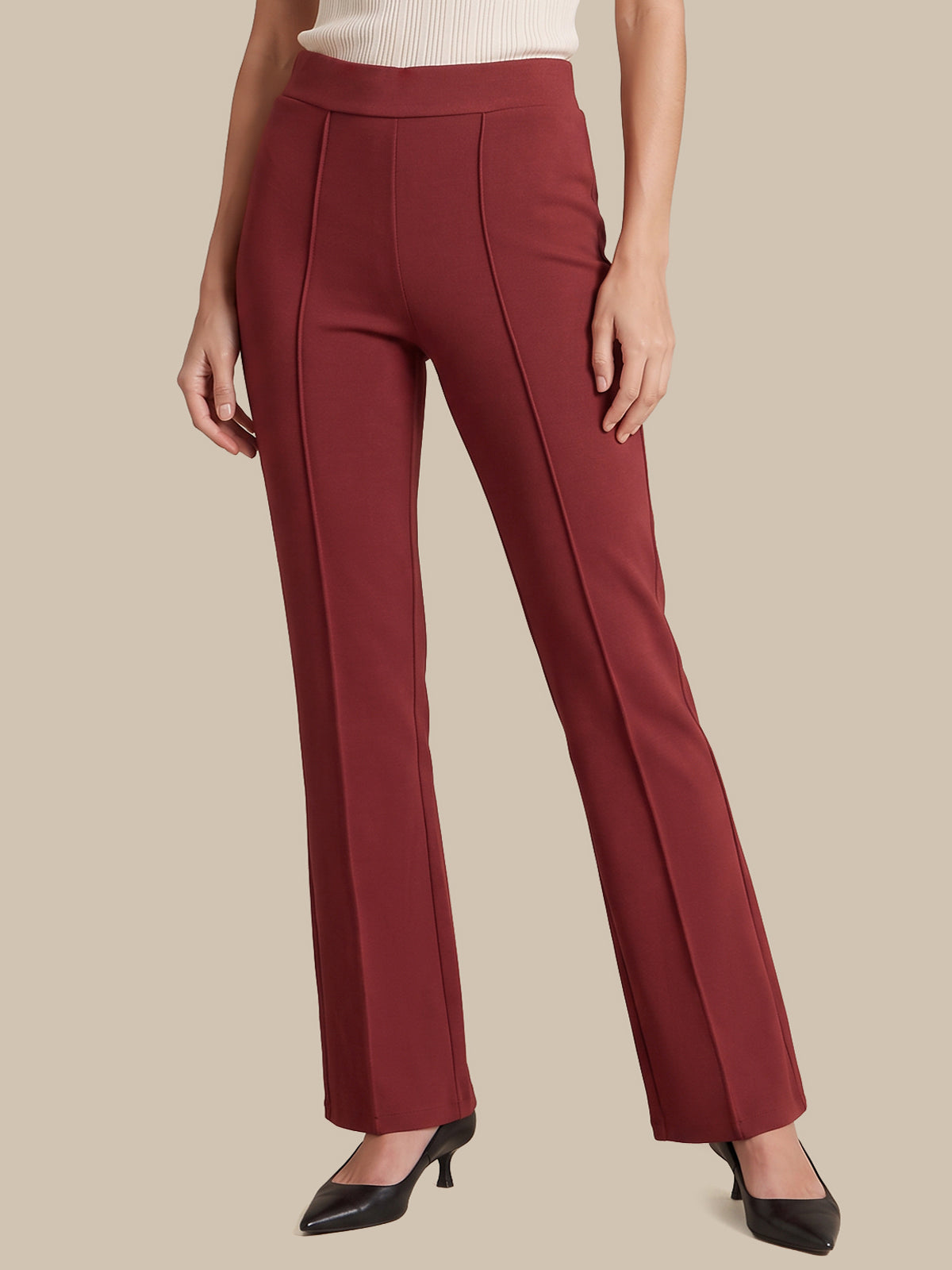A model wearing a pair of high-waisted, straight-leg trousers in a deep burgundy red, paired with a cream ribbed top and black pointed-toe heels, set against a solid beige background.