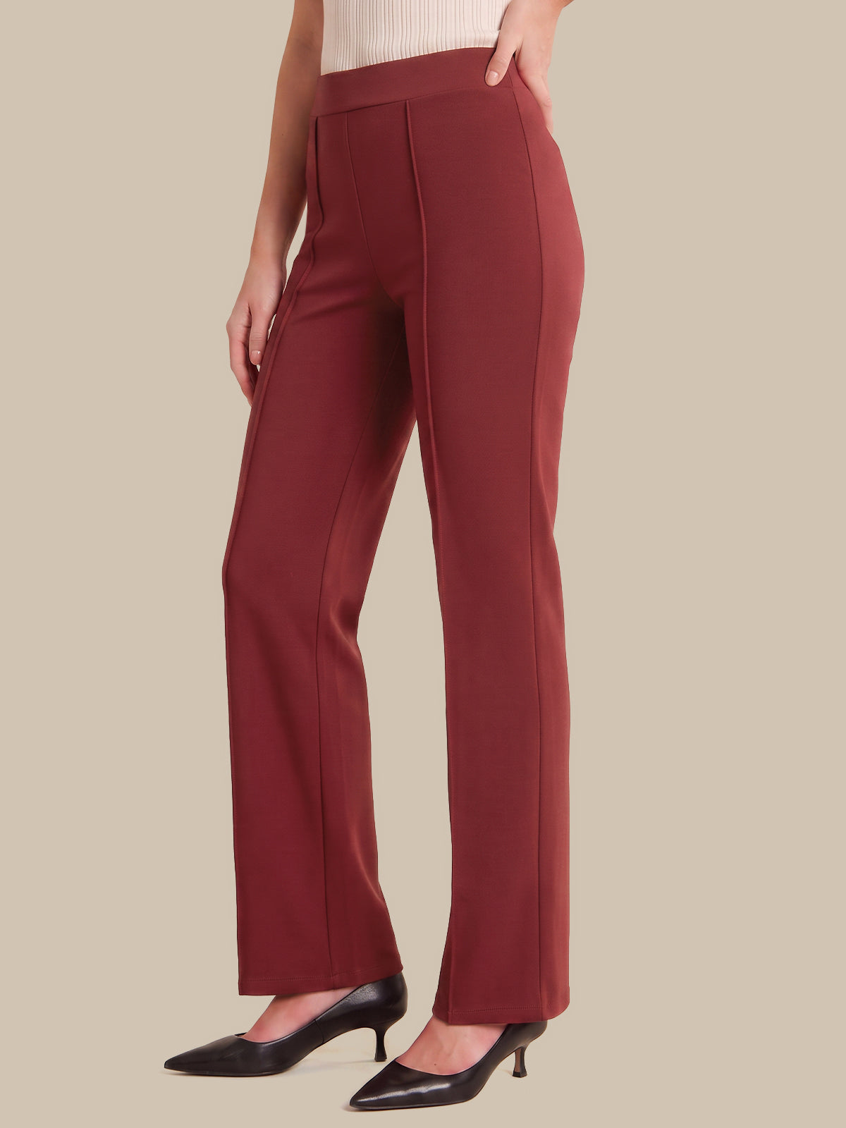 A model wearing a pair of high-waisted, wide-leg trousers in a deep burgundy red, paired with black pointed-toe pumps, shown against a solid beige background.