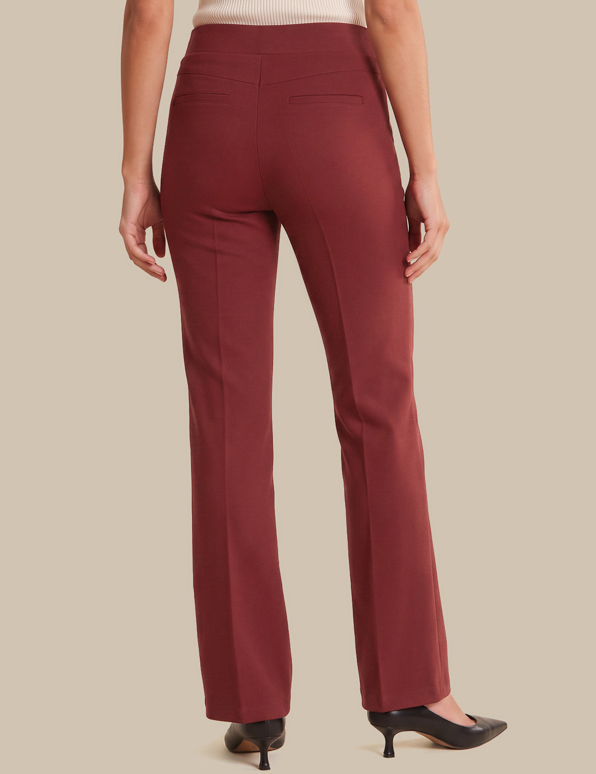 A person shown from the back wearing straight-leg trousers in a deep burgundy red, paired with a cream ribbed top and black pointed-toe heels, against a solid beige background.