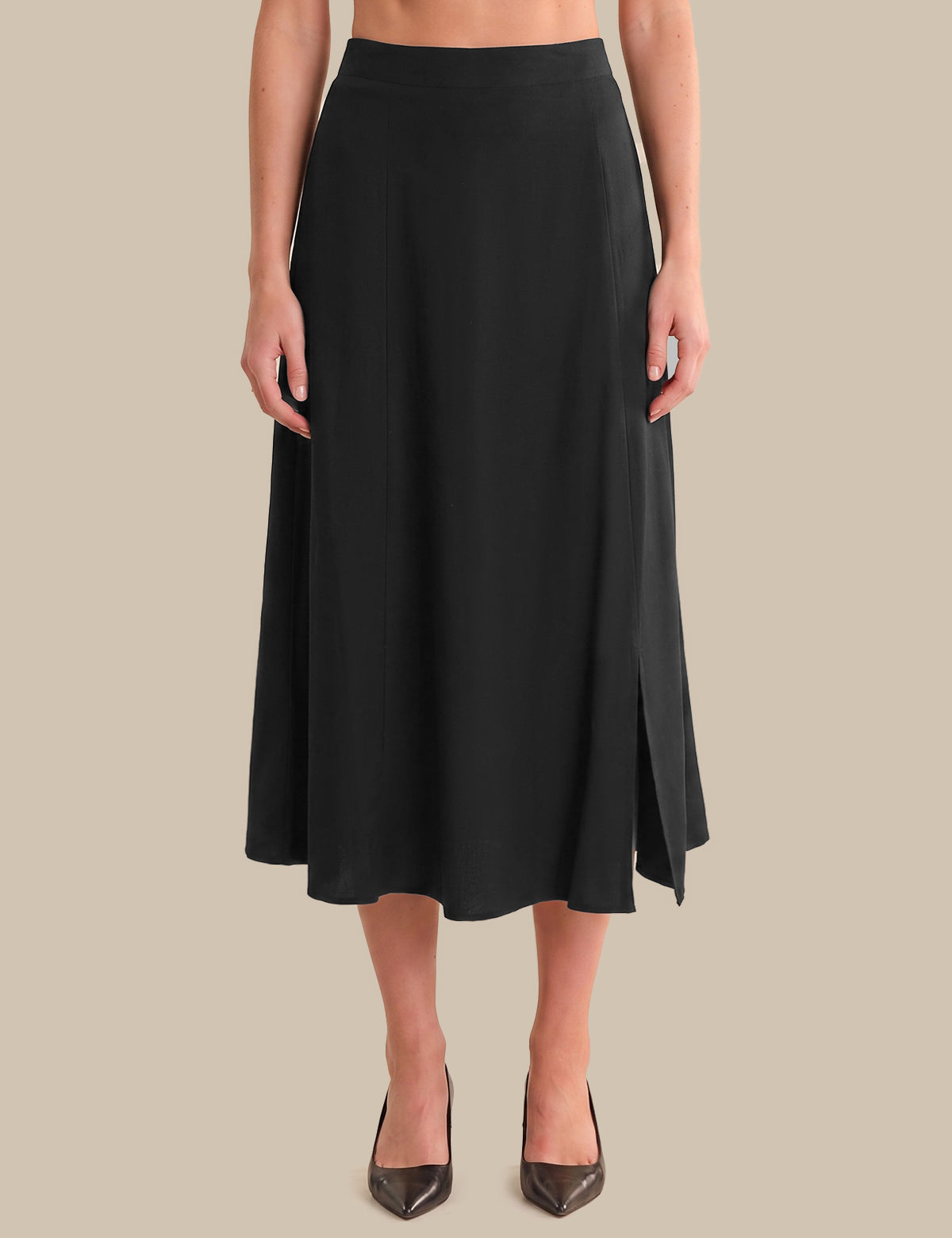 A woman modeling a black midi skirt with side slits, paired with dark brown pointed-toe heels, against a solid beige background.