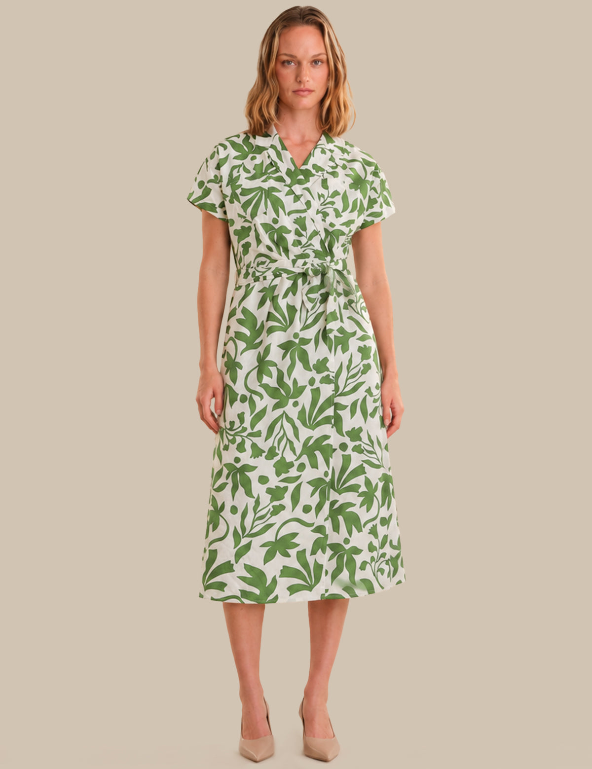 A woman modeling a short-sleeve midi dress with a green botanical leaf print on a white background, standing against a solid beige backdrop.