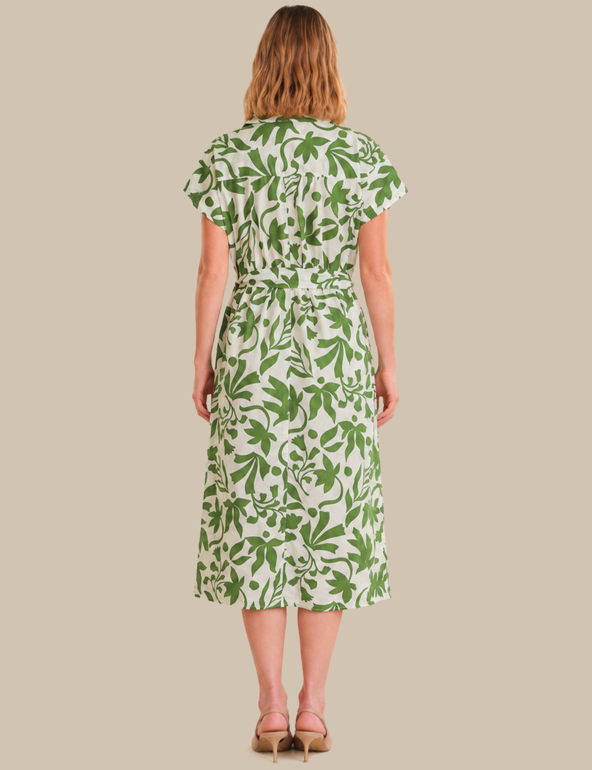 A woman viewed from the back wearing a short-sleeve midi dress with a green botanical leaf pattern on a white background, paired with tan heeled sandals, set against a solid beige studio backdrop.