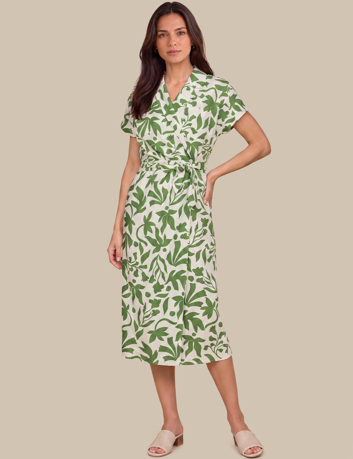 A woman modeling a short-sleeve wrap midi dress with a green leaf pattern on a cream background, paired with beige slide sandals, set against a solid beige studio background.