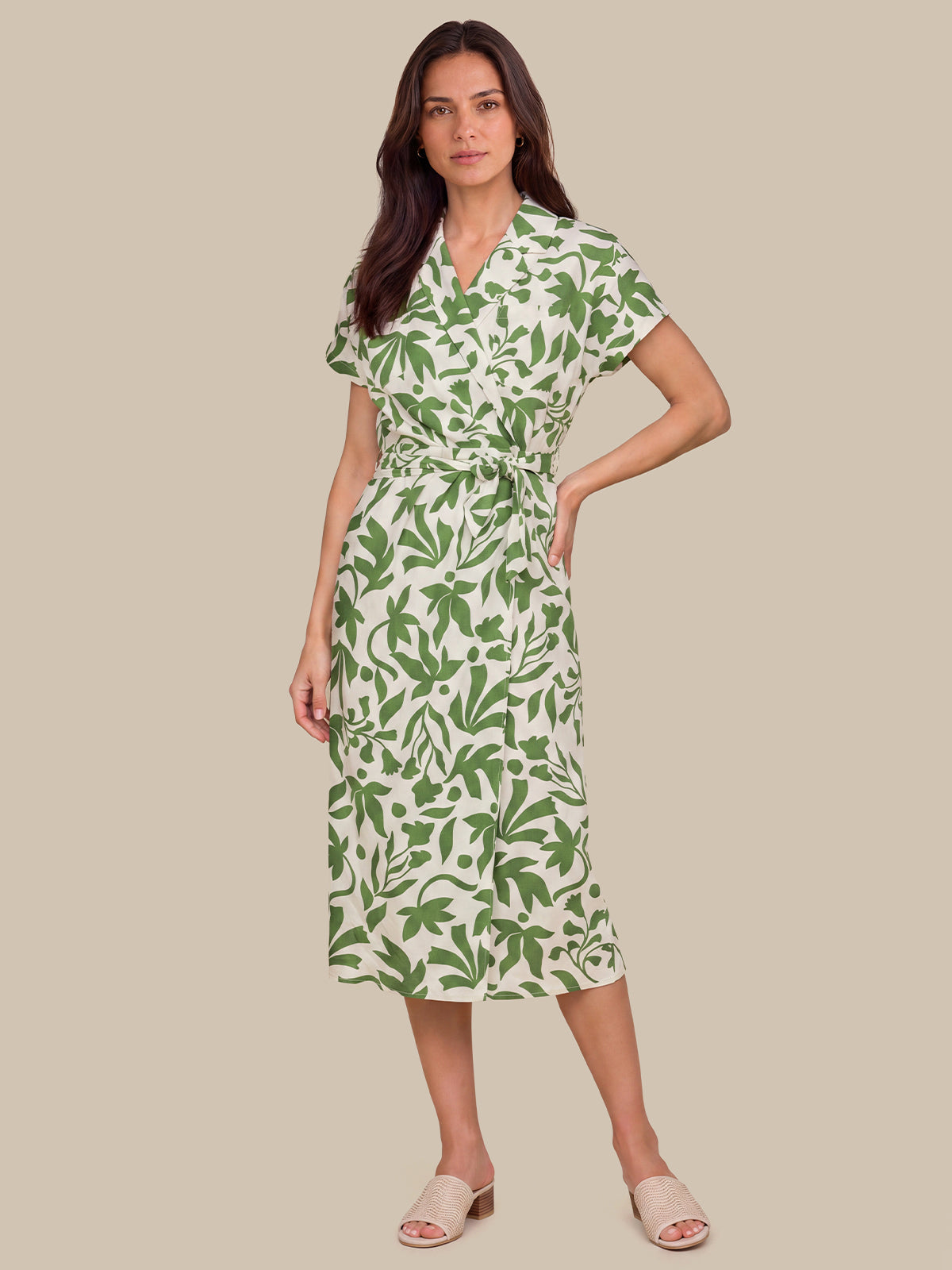 A woman modeling a short-sleeve wrap midi dress with a green leaf pattern on a cream background, paired with beige slide sandals, set against a solid beige studio background.