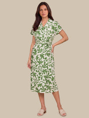 A woman modeling a short-sleeve wrap midi dress with a green leaf pattern on a cream background, paired with beige slide sandals, set against a solid beige studio background.