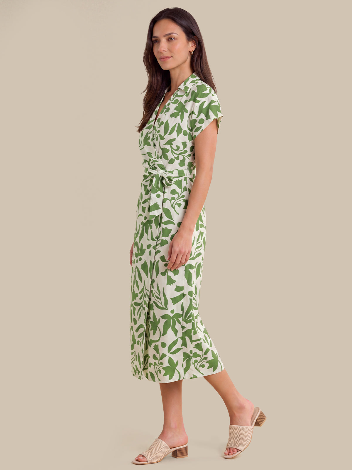 A woman modeling a short-sleeve wrap midi dress with a green leaf pattern on a cream background, paired with beige slide sandals, set against a solid beige studio background.