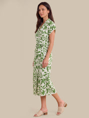 A woman modeling a short-sleeve wrap midi dress with a green leaf pattern on a cream background, paired with beige slide sandals, set against a solid beige studio background.
