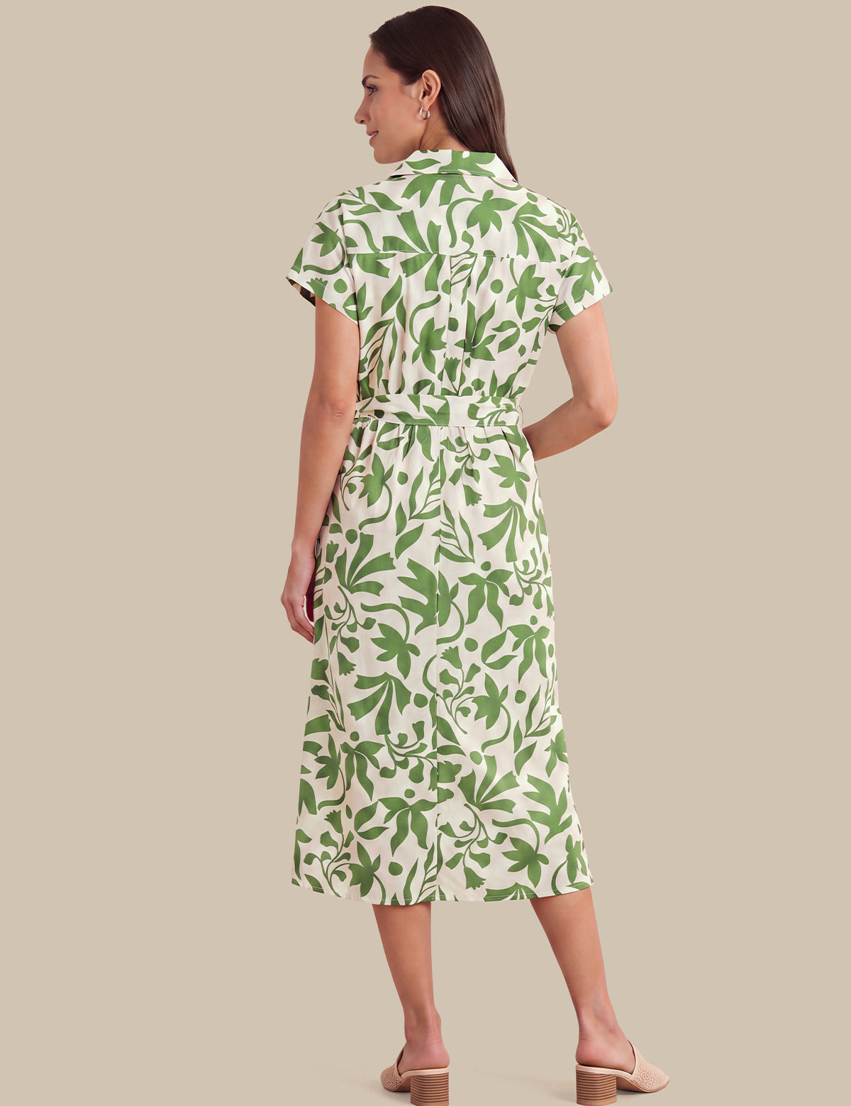 A woman shown from the back wearing a short-sleeve midi dress with a green leaf pattern on a cream background, paired with beige block-heel mule sandals, against a solid beige studio background.