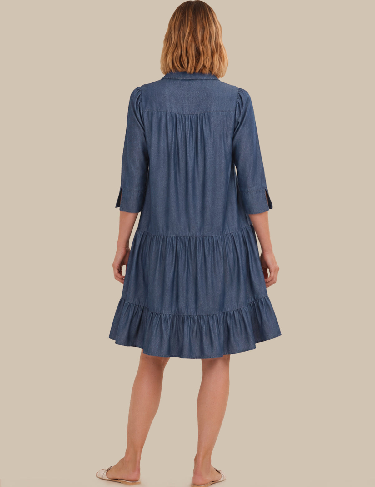 A woman viewed from the back wearing a blue tiered shirt dress with three-quarter sleeves, standing against a solid beige background.
