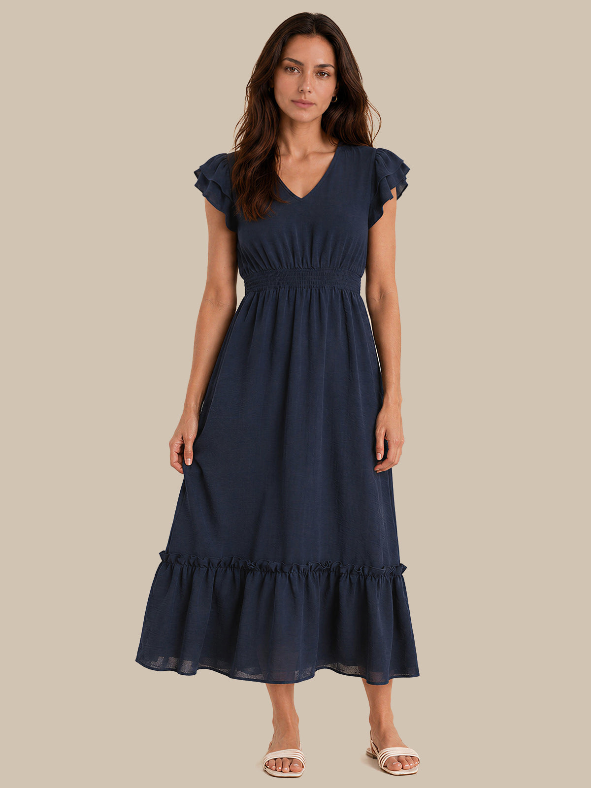 A woman modeling a navy blue midi dress with ruffled cap sleeves, a V-neckline, a gathered waist, and a tiered ruffle hem, set against a solid beige background.