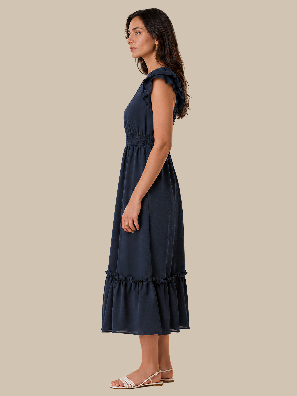 A woman modeling a sleeveless navy blue midi dress with ruffled shoulder details, a gathered waist, and a tiered hem, shown in profile against a solid beige background.