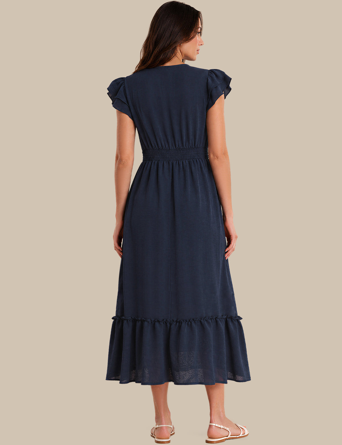 A woman shown from the back wearing a navy blue midi dress with ruffled cap sleeves, a gathered waist, and a ruffled hem, paired with light pink flat sandals against a solid beige background.