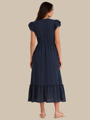 A woman shown from the back wearing a navy blue midi dress with ruffled cap sleeves, a gathered waist, and a ruffled hem, paired with light pink flat sandals against a solid beige background.
