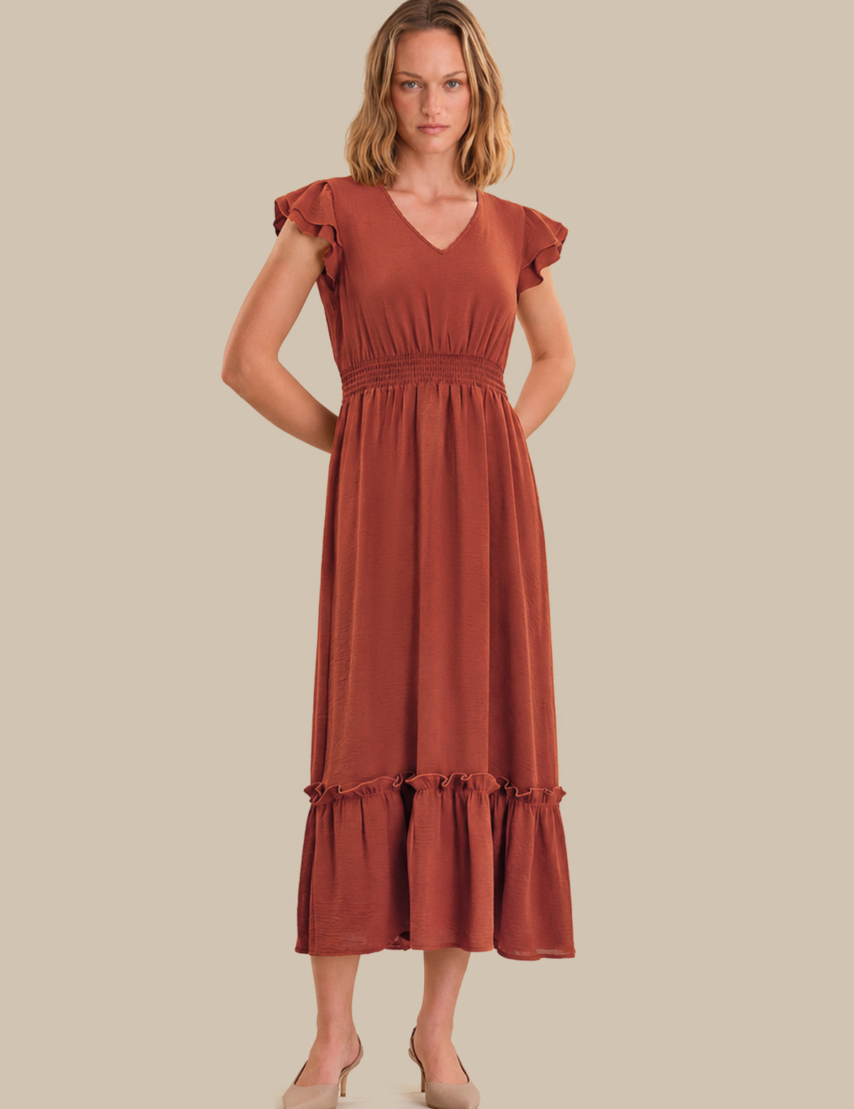 A woman modeling a rust-colored midi dress with ruffled cap sleeves, a V-neckline, a smocked waist, and a tiered ruffle hem, set against a solid beige background.