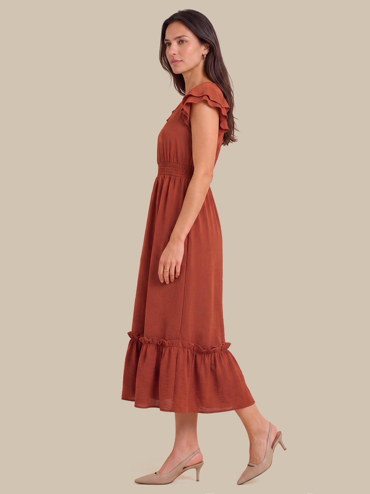 A woman modeling a rust-colored midi dress with ruffled shoulder details and a ruffled hem, paired with beige slingback heels, set against a solid beige background.