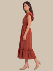 A woman modeling a rust-colored midi dress with ruffled shoulder details and a ruffled hem, paired with beige slingback heels, set against a solid beige background.