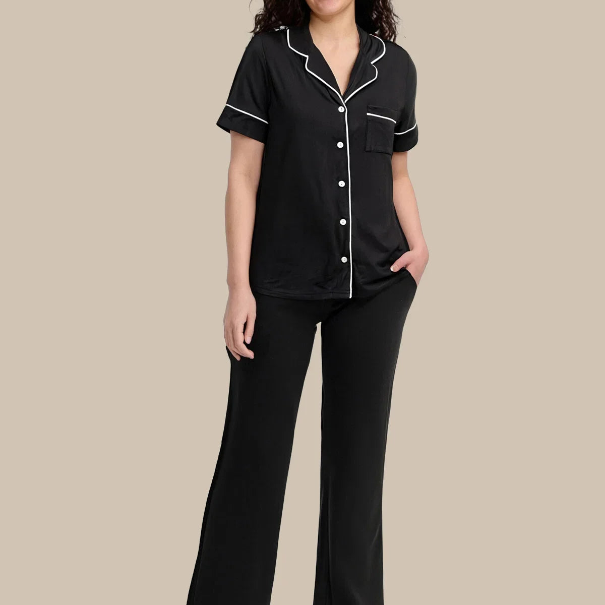 A woman modeling a two-piece black pajama set with white piping details, consisting of a short-sleeve button-up top and matching pants, against a solid beige background.
