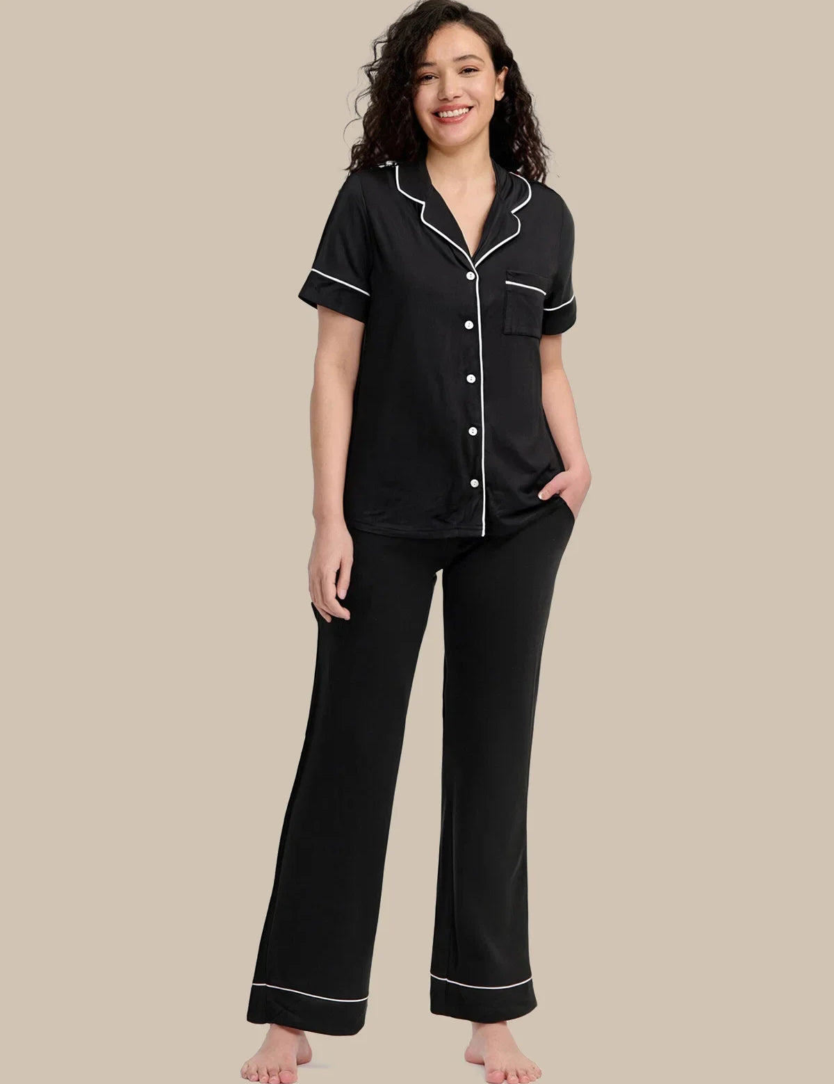 A woman modeling a two-piece black pajama set with white piping details, consisting of a short-sleeve button-up top and matching pants, against a solid beige background.