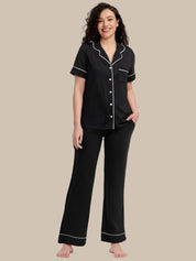 A woman modeling a two-piece black pajama set with white piping details, consisting of a short-sleeve button-up top and matching pants, against a solid beige background.