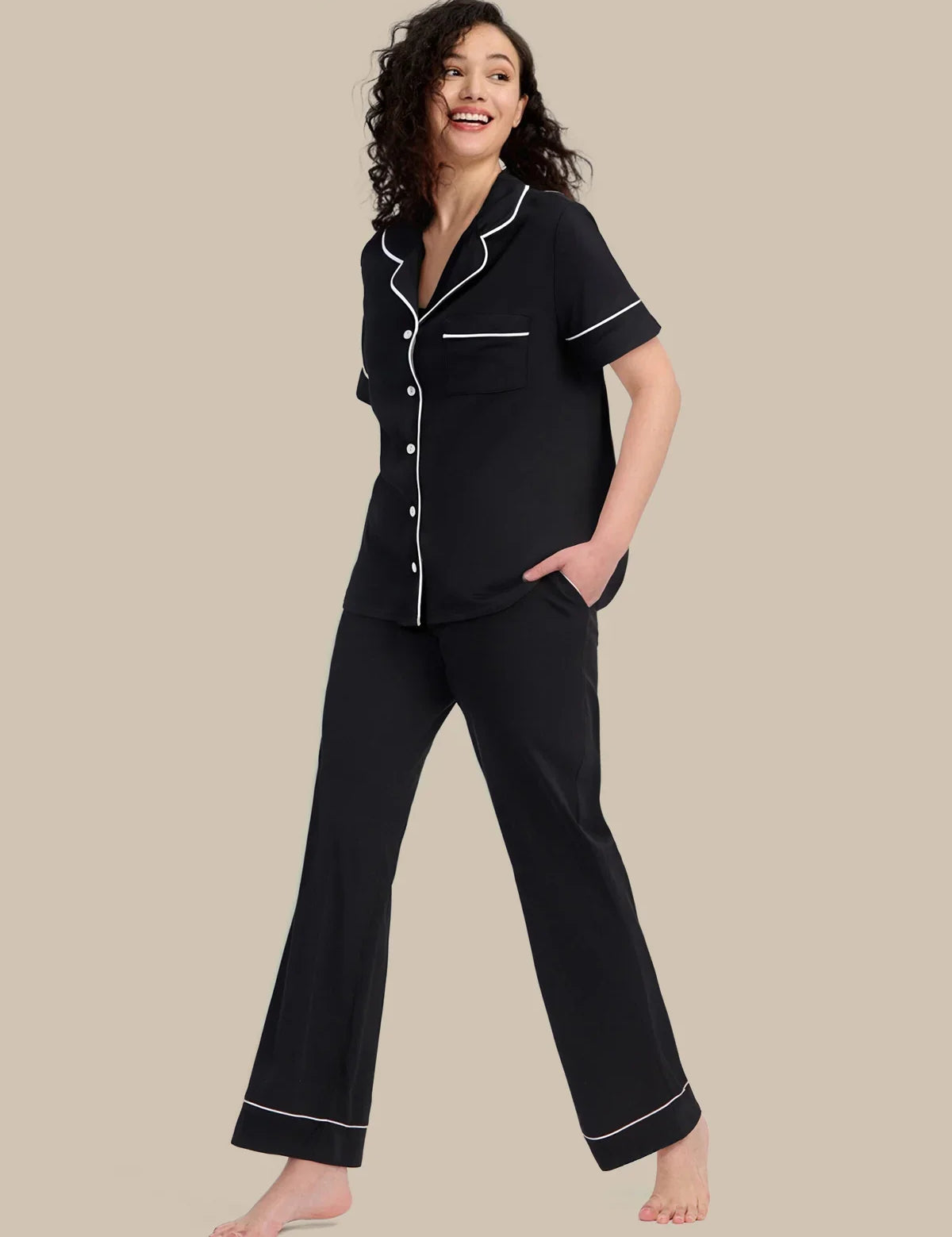 A woman modeling a black short-sleeve pajama set with white piping details, consisting of a button-up top and matching wide-leg pants, against a solid beige background.