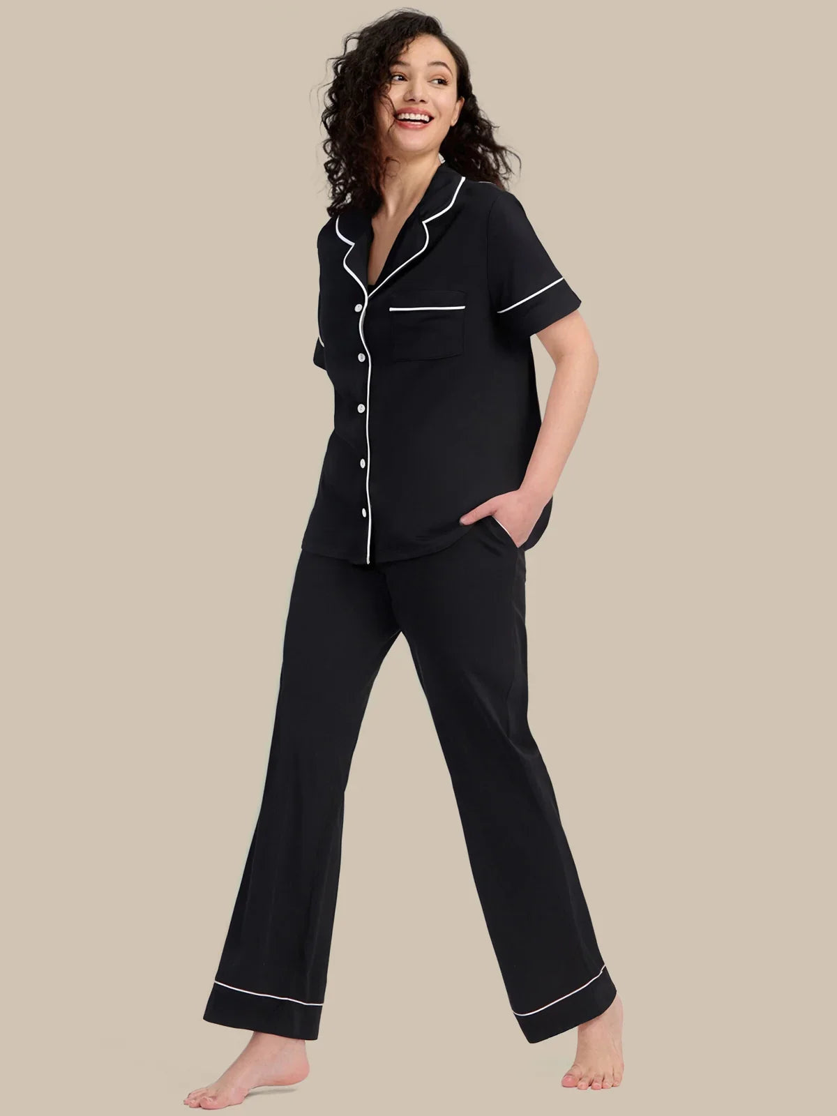 A woman modeling a black short-sleeve pajama set with white piping details, consisting of a button-up top and matching wide-leg pants, against a solid beige background.