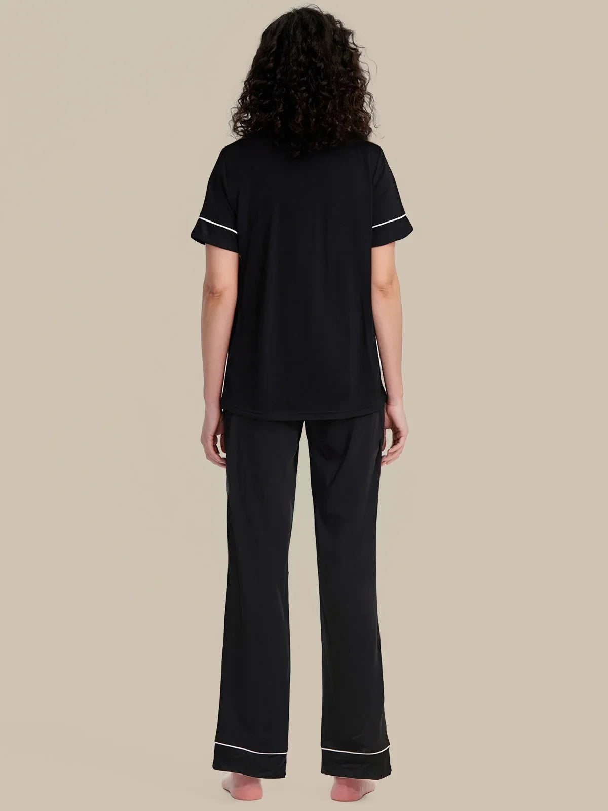 A woman shown from the back wearing a black short-sleeve pajama top and matching wide-leg pajama pants with thin white piping trim, set against a solid beige background.