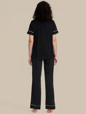 A woman shown from the back wearing a black short-sleeve pajama top and matching wide-leg pajama pants with thin white piping trim, set against a solid beige background.