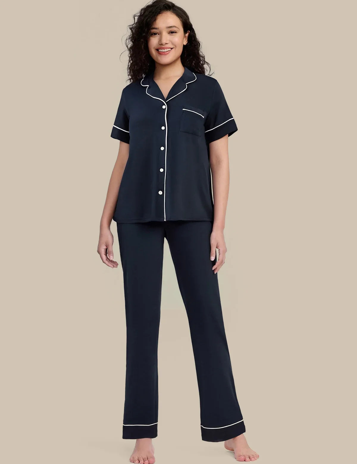 A woman modeling a two-piece navy blue pajama set with white piping details, consisting of a short-sleeve button-up top and matching pants, against a solid beige background.