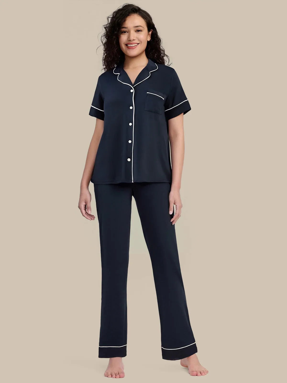 A woman modeling a two-piece navy blue pajama set with white piping details, consisting of a short-sleeve button-up top and matching pants, against a solid beige background.