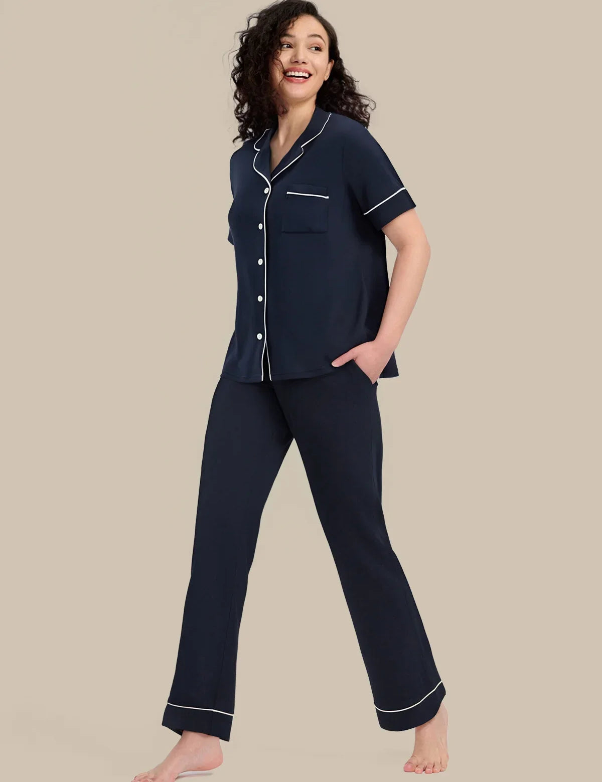 A woman modeling a navy blue pajama set with white piping details, consisting of a short-sleeve button-up top and matching pants, against a solid beige background.