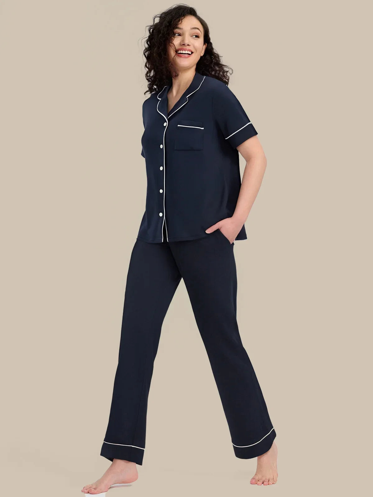 A woman modeling a navy blue pajama set with white piping details, consisting of a short-sleeve button-up top and matching pants, against a solid beige background.