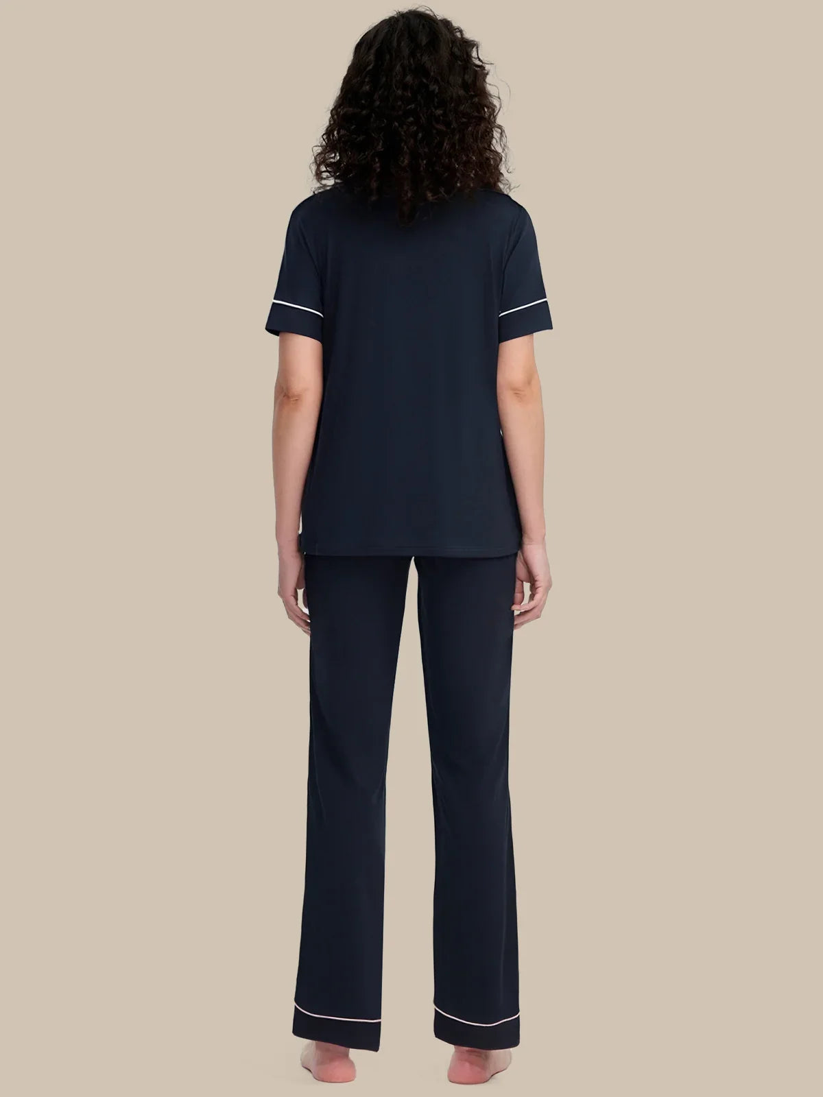 A woman viewed from the back wearing a navy blue short-sleeve pajama top and matching wide-leg pajama pants with thin white piping trim on the sleeve cuffs and hems, set against a solid beige background.